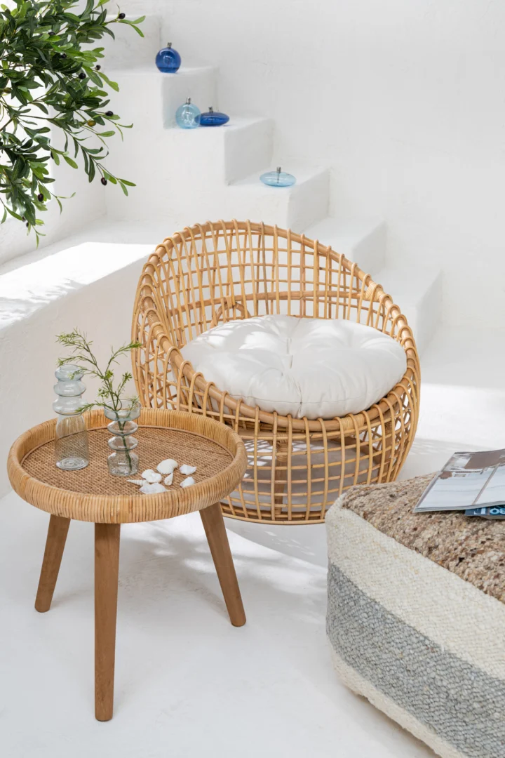 Een gezellige ronde rotan stoel met een wit kussen staat naast de Set van 2 bijzettafels Lurne Rotan/Sparrenhout Natuurlijk, waarin glazen vazen en schelpen staan. Op de achtergrond staan een witte trap, blauw glasdecor en groene plantenbladeren.