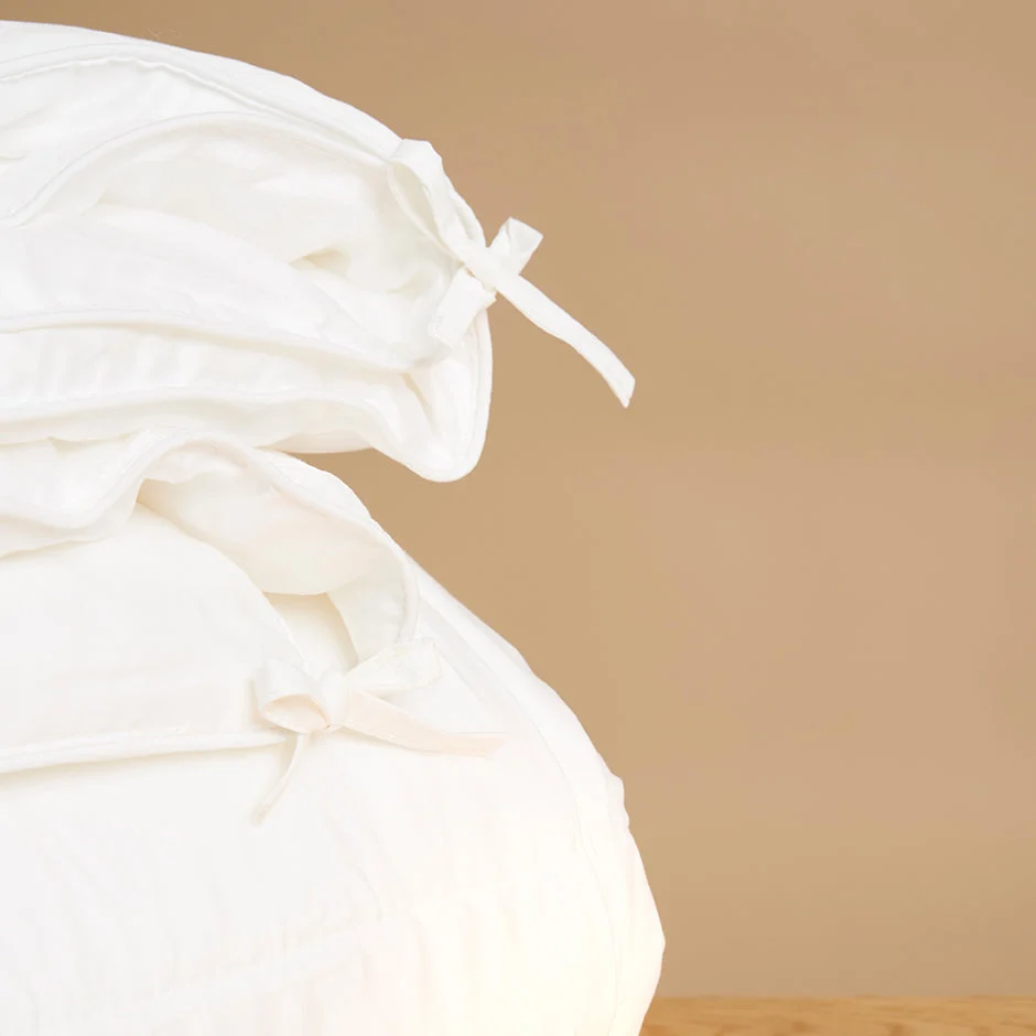 Een close-up van de Dekbedovertrek 100x135 Ontspannen sfeer toont wit beddengoed met strikken van stof tegen een effen beige achtergrond.