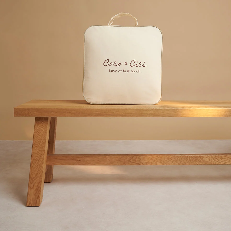 Een crèmekleurige vierkante tas met het label "Dekbedovertrek 100x135 Ontspannen sfeer" met een handvat rust op een eenvoudige houten bank tegen een beige achtergrond.