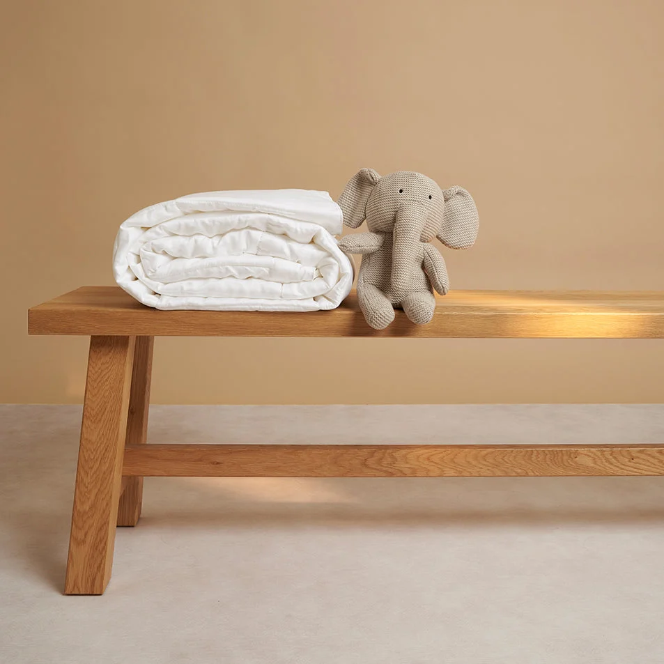 De Dekbedovertrek 100x135 Ontspannen sfeer ligt netjes opgevouwen op een houten bankje, naast een grijs gebreid olifantenpluche, beide tegen een beige achtergrond.