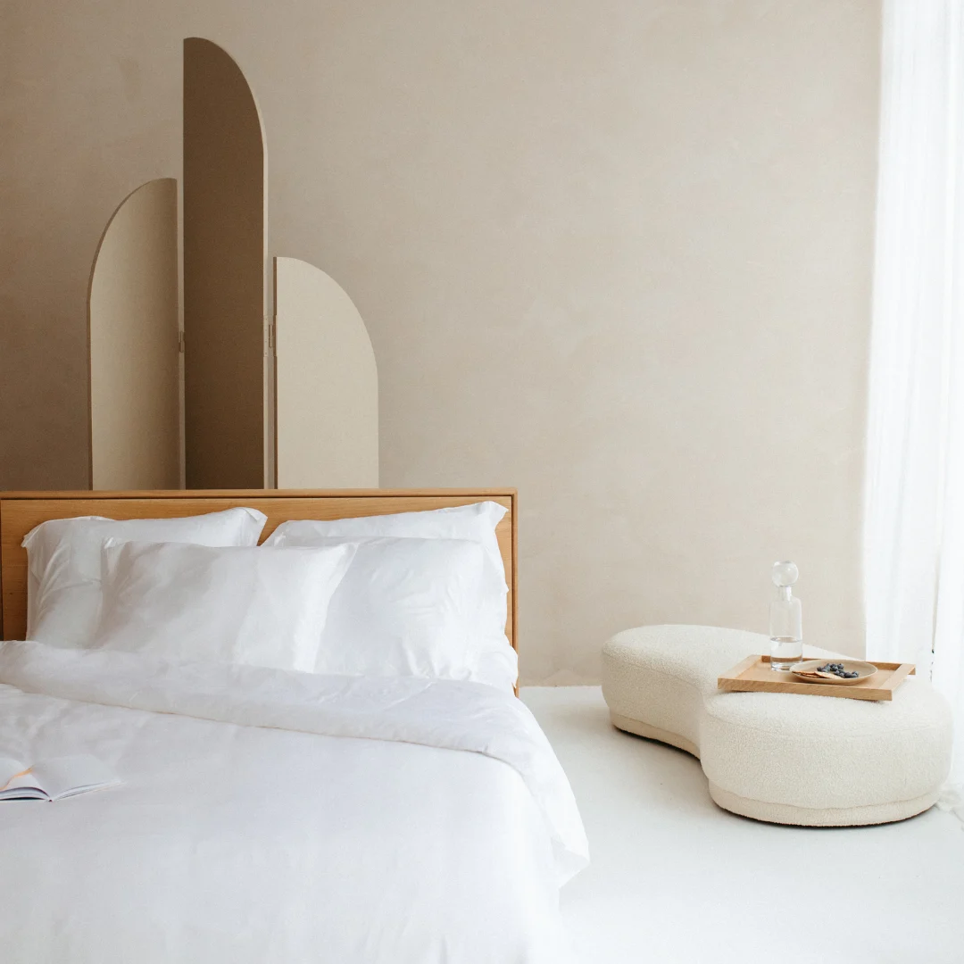 Een minimalistische slaapkamer met het Dekbed Koningin (200 x 220) Comfortabel klimaat op een netjes opgemaakt bed met wit linnengoed, een houten hoofdeinde, beige scheidingswand, sculpturale witte voetenbank met dienblad en transparante gordijnen die natuurlijk licht binnenlaten.