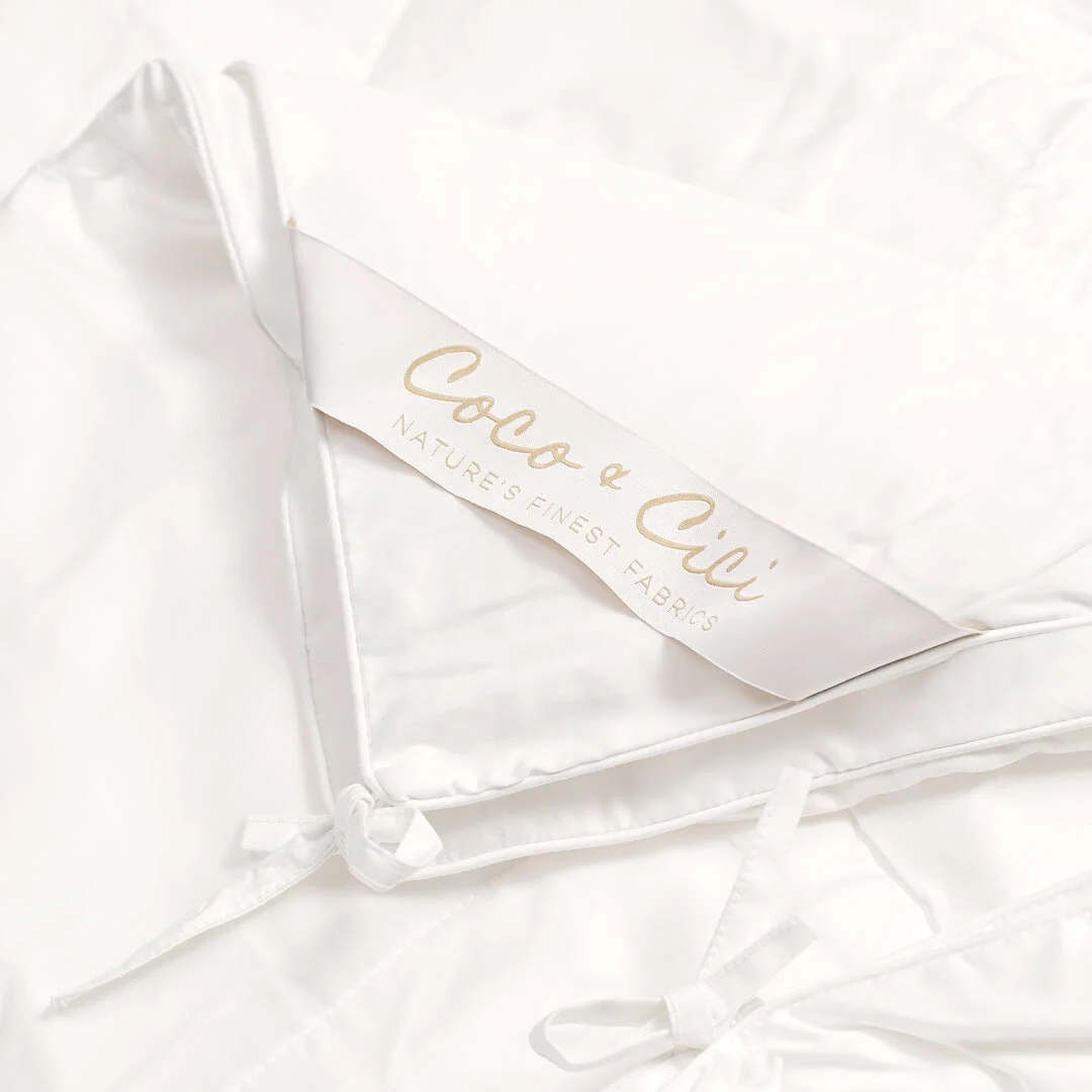 Een close-up van de Dekbed Koningin (200 x 220) Comfortabel klimaat toont de witte stof, gladde zijdeachtige textuur, bandjes en een label met gouden "Coco & Cici Nature's Finest Fabrics" letters.