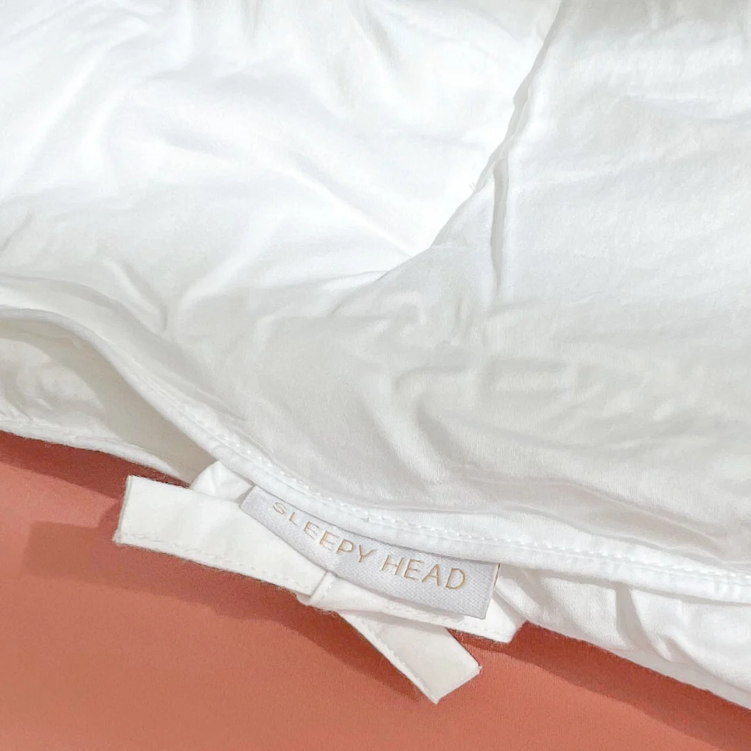 Een close-up van het Dekbed Koningin (200 x 220) Comfortabel klimaat, een wit en donzig kussen met een stoffen label, rustend op een roze ondergrond. Het kussen ziet er zacht en uitnodigend uit.