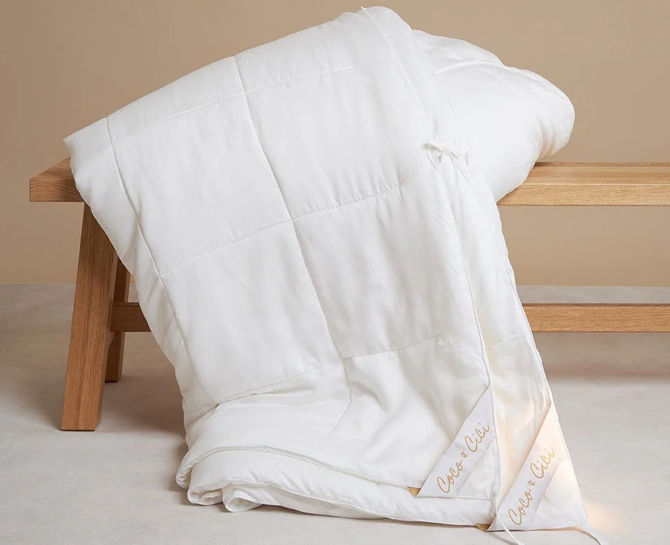 Een Dekbed 240x220 Oase van roest gedrapeerd over een eenvoudige houten bank op een lichte vloer, met zijn witte, gewatteerde stof en subtiele borduursel in de hoek.