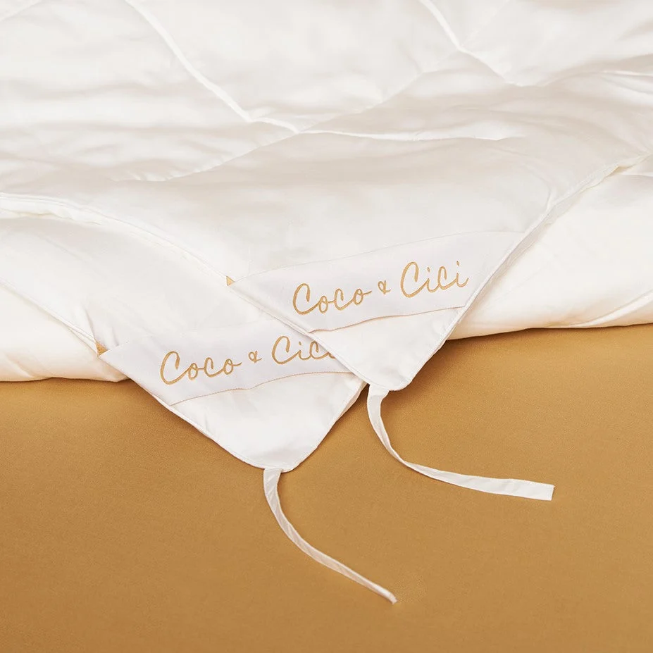 Het Dekbed 240x220 Oase van rust is een wit, doorgestikt dekbed met goudkleurig "Coco & Cici" schrift in de hoek, liggend op een gebruind bed en met zichtbare witte hoekbanden.