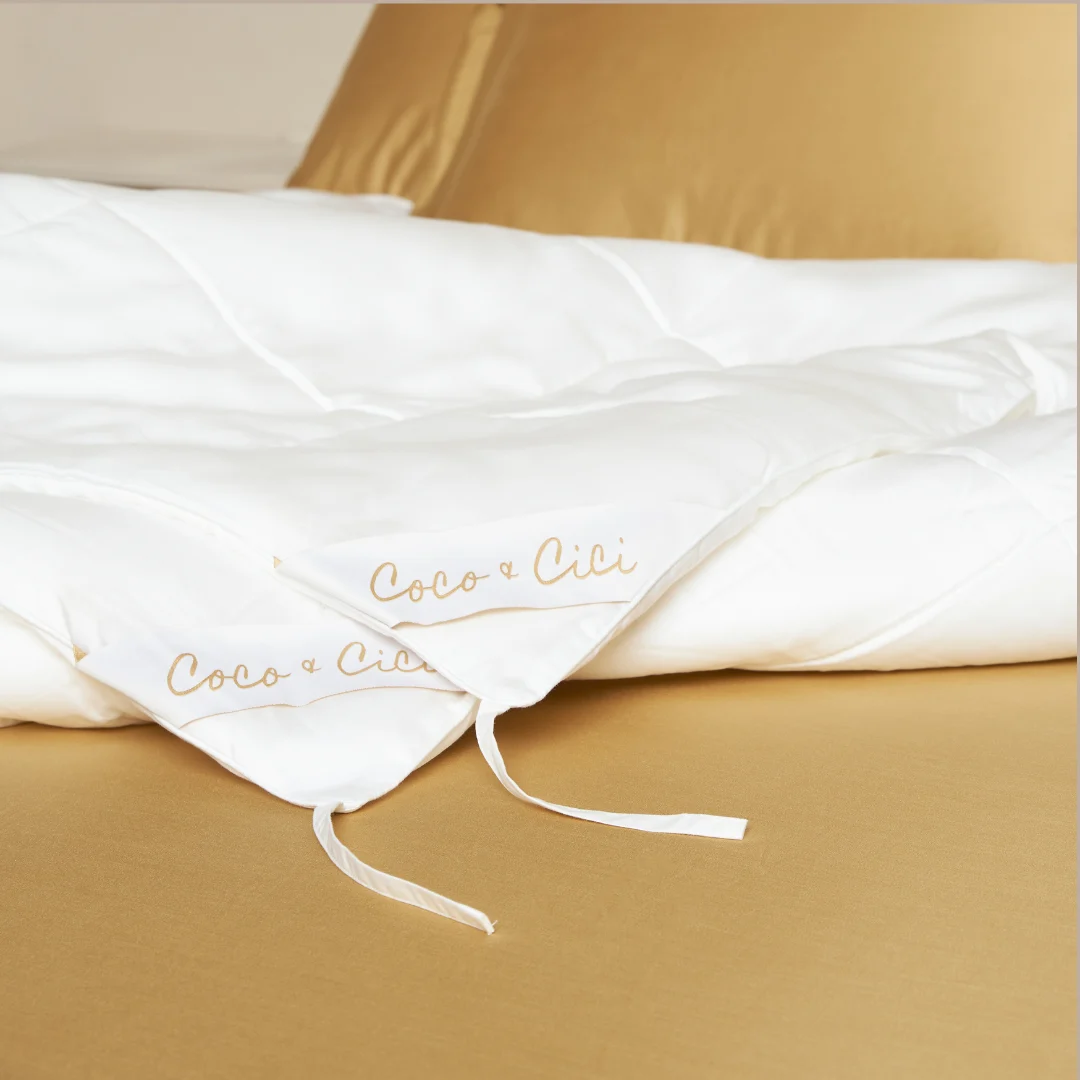 Het Dekbed 240x220 Stijlvolle upgrade, met Coco & Cici geborduurd in goud en witte stoffen banden op de hoeken, rust op een netjes opgemaakt bed met bruine lakens.