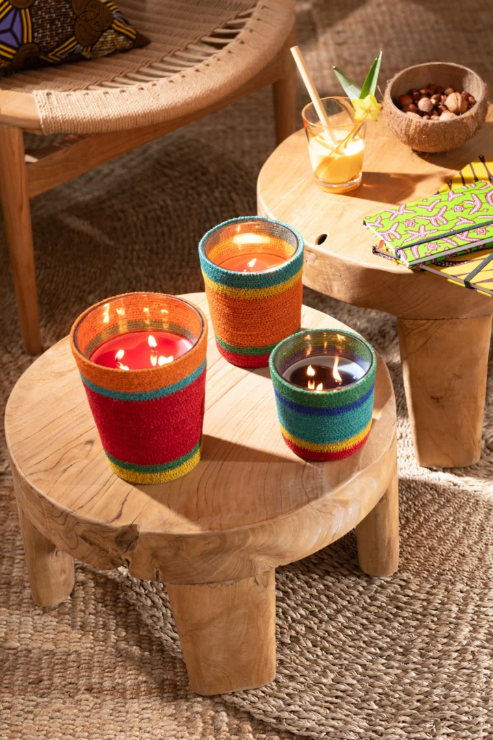 Drie kleurrijke kaarsen met kralen branden op de ronde teakhouten tafel van Bijzettafel Roest, naast een drankje met een rietje, een schaaltje nootjes en servetten met een patroon. Vlakbij staan een geweven stoel en een gevlochten kleed.