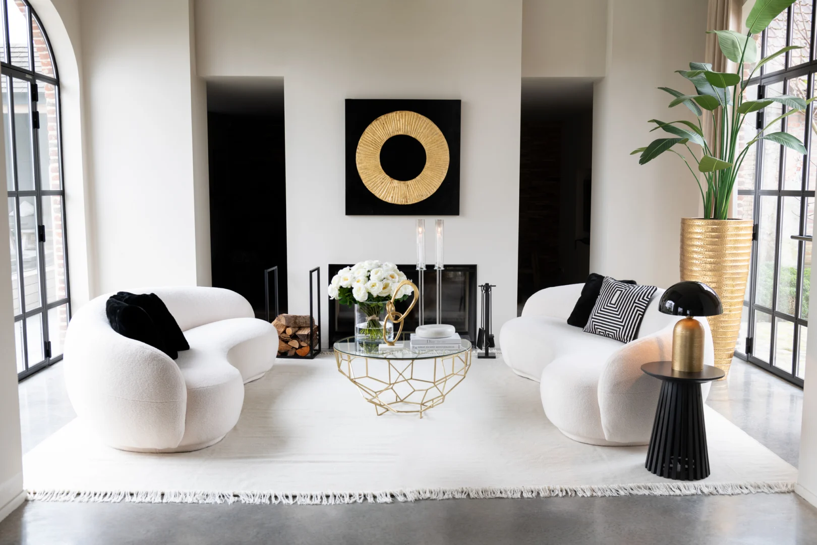 Moderne woonkamer met twee witte gebogen banken, de Salontafel Goud 90 x 90 CM Vierkant metaal salontafel, zwarte en gouden decoraccenten, een grote kamerplant en ramen van vloer tot plafond die de ruimte overspoelen met licht.