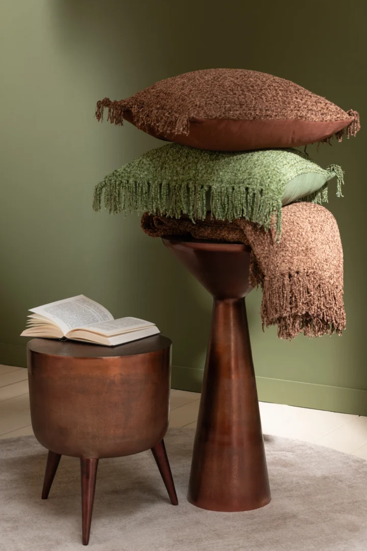 Een vierkante aluminium Bijzettafel Brons (34 x 41 CM) houdt een boek open naast een hoge houten sokkel met drie bruine en groene kussens met kwastjes, tegen een groene muur en een zacht tapijt.