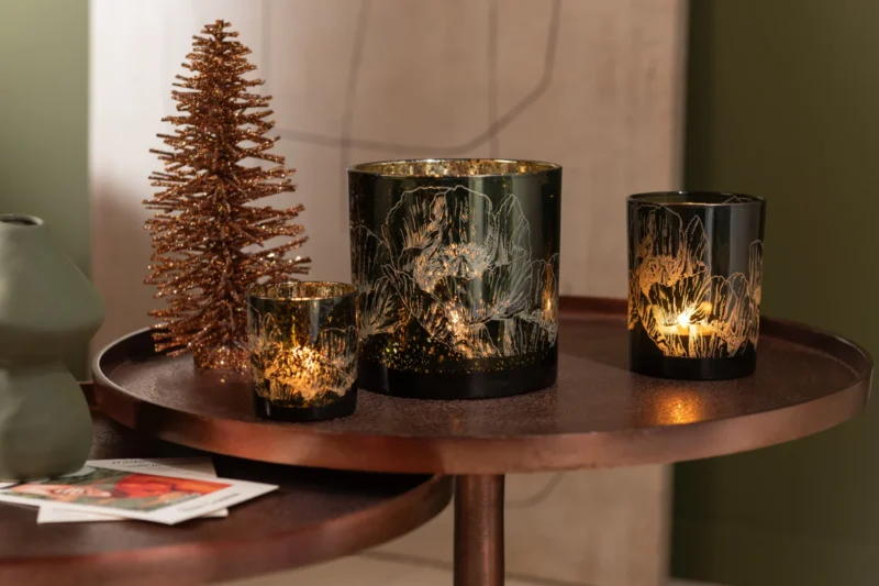 Twee decoratieve kandelaars met gouden bloemmotieven staan op de Set van 2 tafels Aluminium Brons naast een metalen kerstboom en groene vaas, waardoor een gezellige en feestelijke display ontstaat.