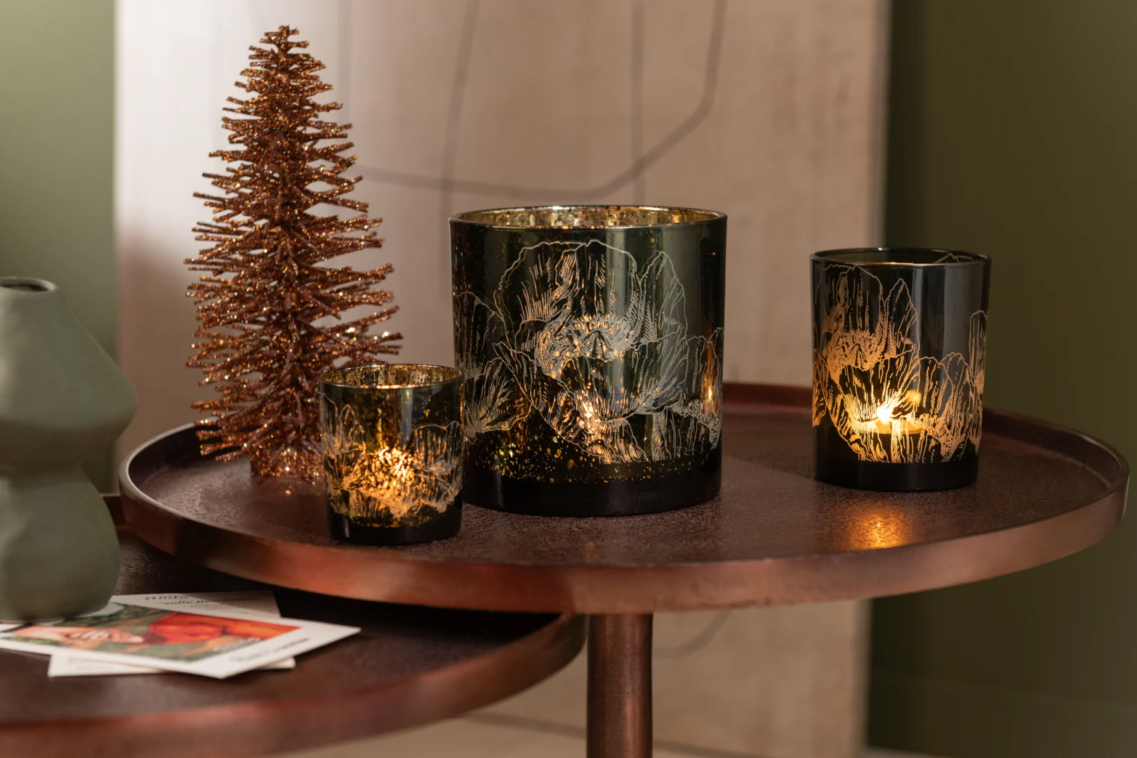 Twee decoratieve kandelaars met gouden bloemmotieven staan op de Set van 2 tafels Aluminium Brons naast een metalen kerstboom en groene vaas, waardoor een gezellige en feestelijke display ontstaat.