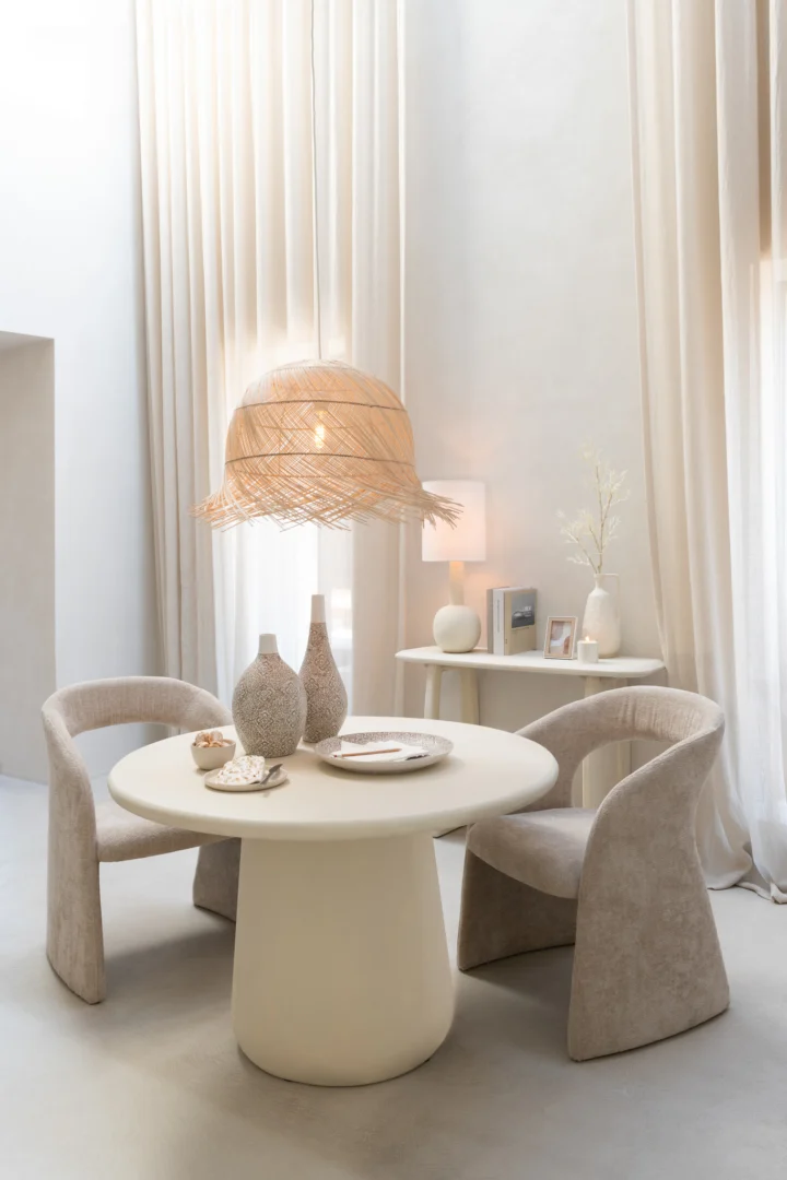 Een moderne eethoek heeft twee beige stoelen en de Eettafel Ram Berkenmultiplex Beige, een ronde crèmekleurige tafel versierd met vazen en borden. Gordijnen, een rieten hanglamp en een consoletafel maken de minimalistische look compleet.