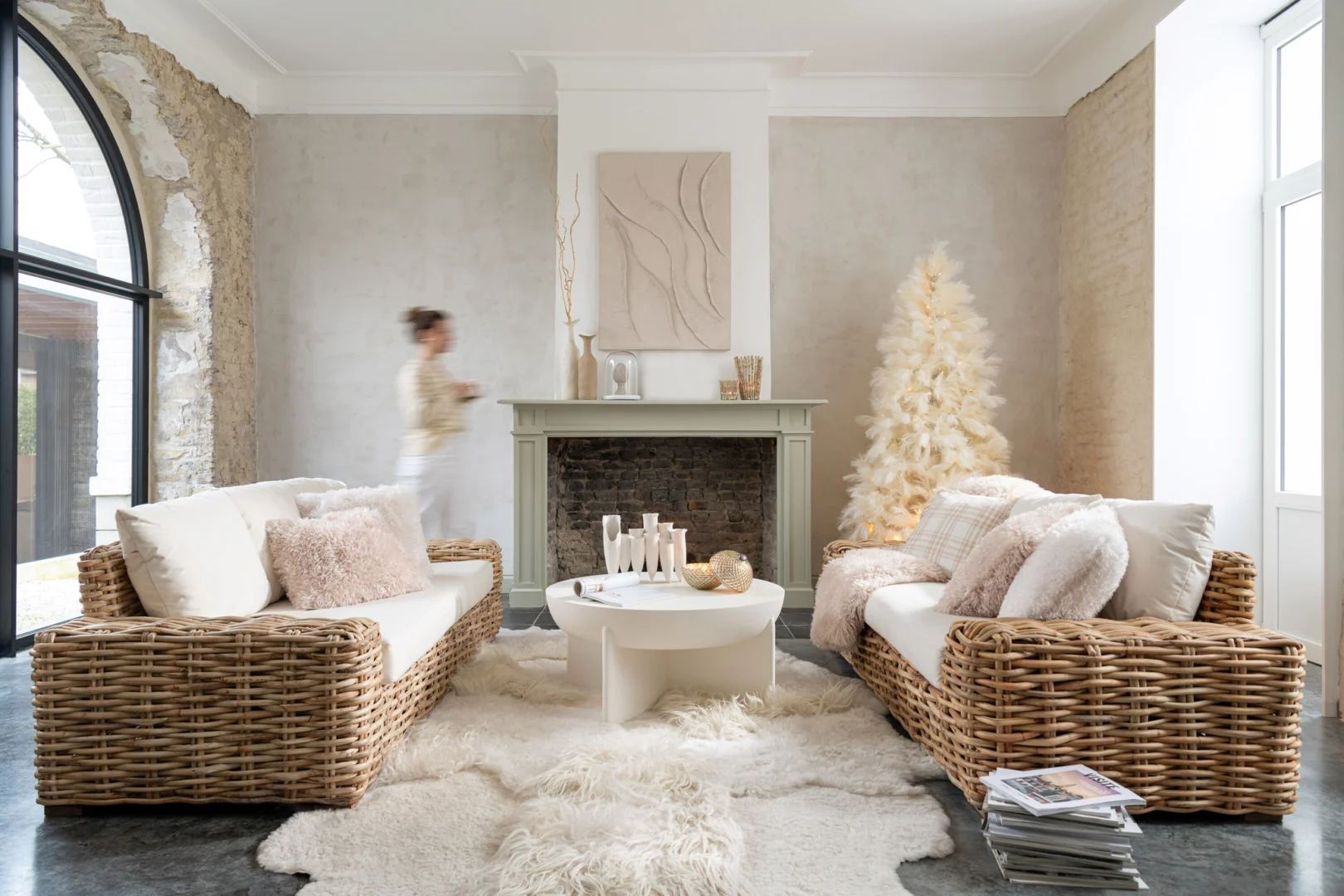 Een lichte, gezellige woonkamer met rieten banken, witte kussens en een donzig vloerkleed is voorzien van een Salontafel Beige, Roest 90 x 90 CM-een elegante vierkante salontafel gemaakt van mangohout. Een witte decoratieve boom en moderne kunst sieren de open haard als er iemand langsloopt.