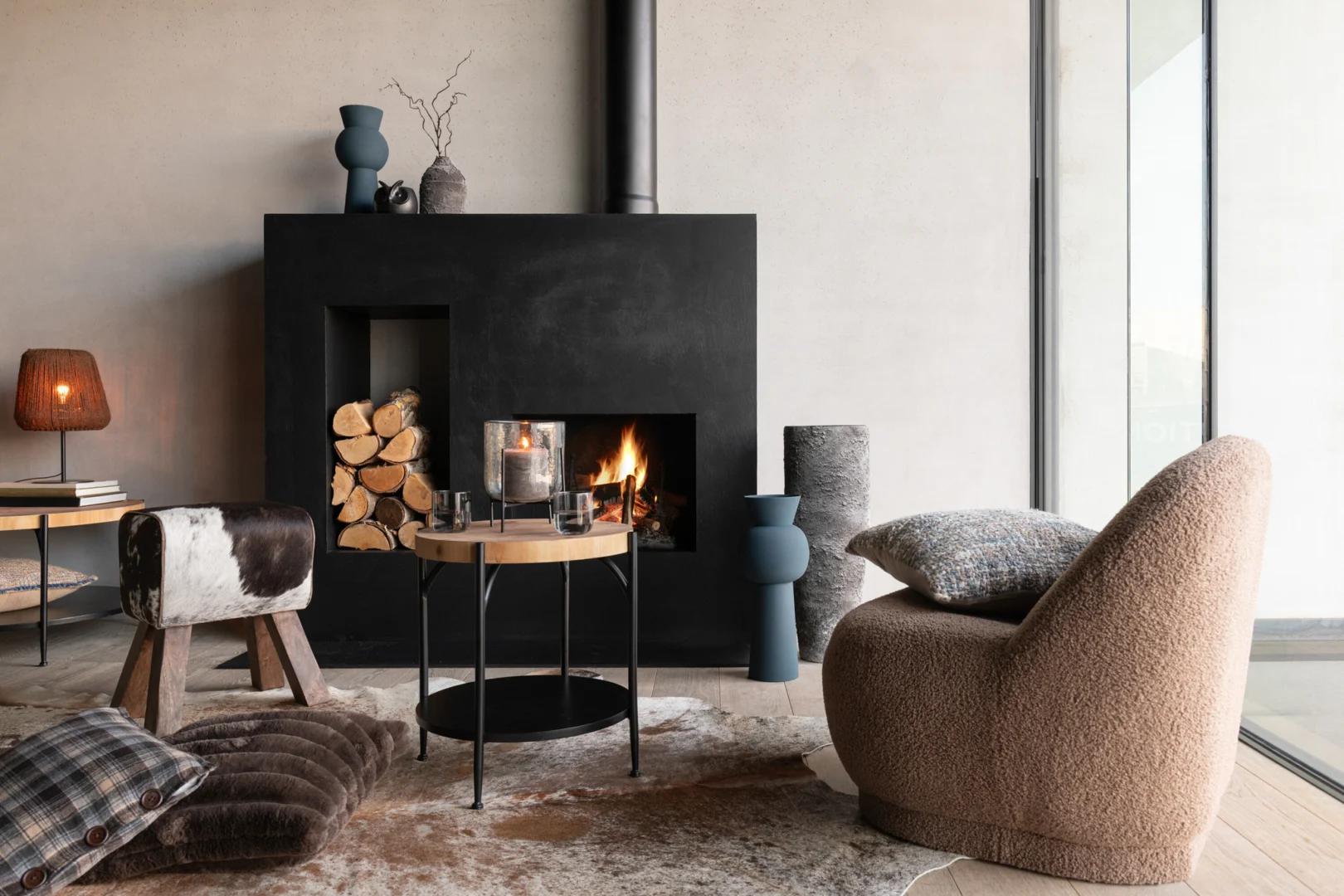 Moderne woonkamer met de Bijzettafel Bio Mdf/IJzer Zwart/Natuurlijk als ronde salontafel, zwarte open haard, gestapeld haardhout, stoelen met structuur, zachte kussens en een groot raam. Warme neutrale tinten versterken de gezellige sfeer.