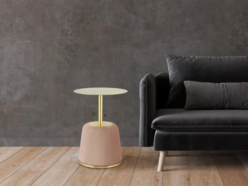 Een moderne woonkamer met een donkergrijze fluwelen bank en de Bijzettafel Roze Rond Ijzer Scandinavisch - een ronde bijzettafel met een gouden blad en een blush roze onderstel - tegen een donkere muur met textuur en een lichte houten vloer.