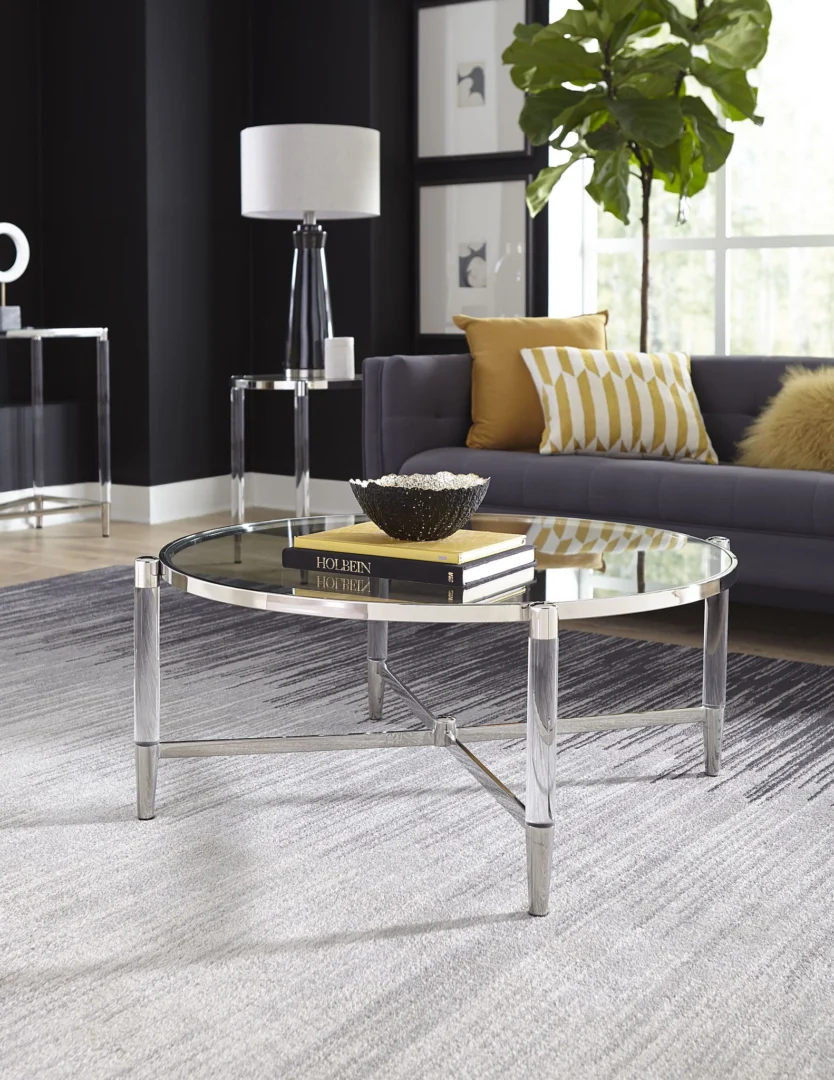 In een moderne woonkamer staat de Salontafel Rond Glas met boeken en een decoratieve schaal, een grijze bank met gele en witte kussens, een staande lamp, een grote plant en muurkunst bij het raam.