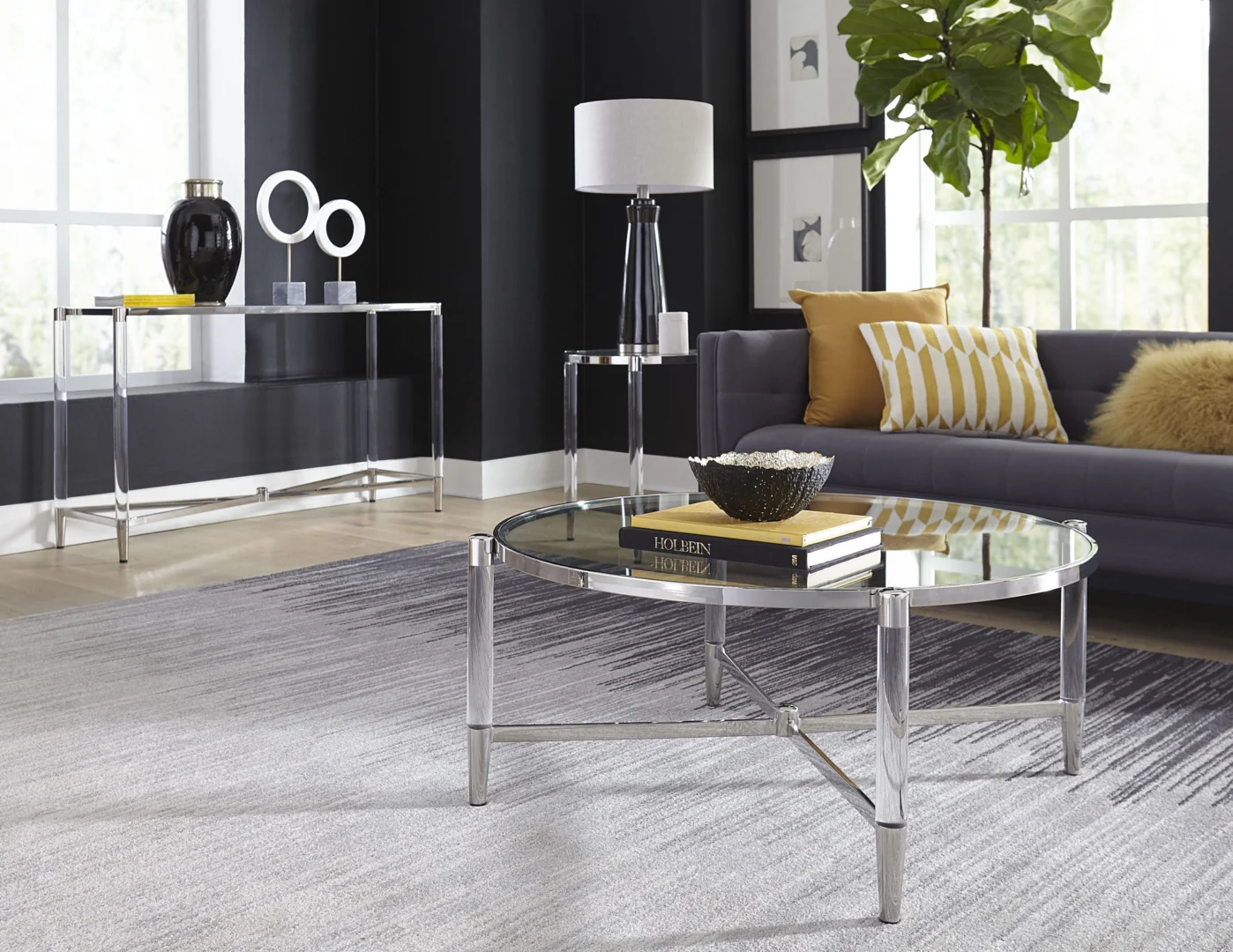 Een moderne woonkamer met de Salontafel Rond Glas salontafel, een grijze bank met gele en witte kussens, een hoge lamp, een potplant, levendige kunstwerken en grote ramen die natuurlijk licht binnenlaten.