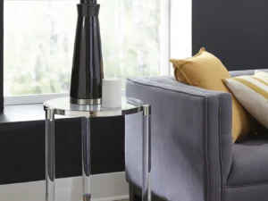 Een moderne woonkamer met een grijze bank, gele en witte kussens, een Marilyn Glas, Acryl en Gepolijst Roestvrijstalen Bijzettafel, een zwart met zilveren lamp met een witte kap en een klein wit decoratief object.