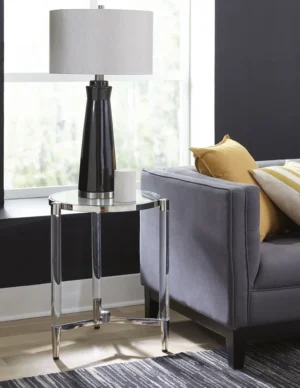 Een moderne woonkamer met een grijze bank, gele en witte kussens, een Marilyn Glas, Acryl en Gepolijst Roestvrijstalen Bijzettafel, een zwart met zilveren lamp met een witte kap en een klein wit decoratief object.