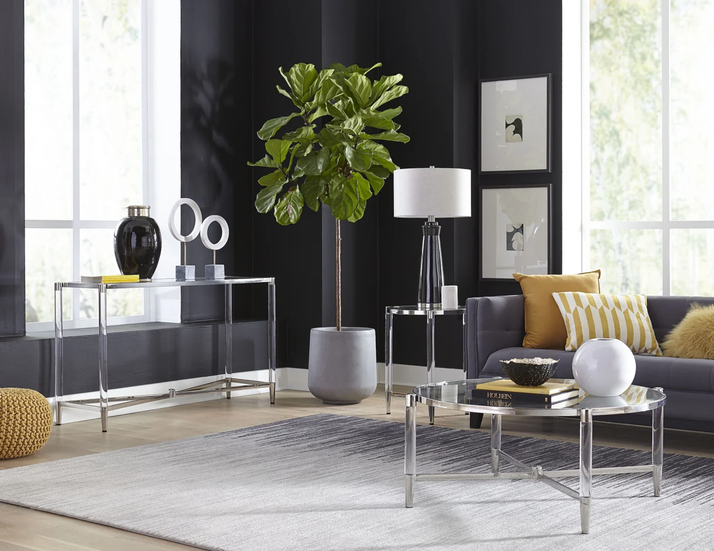 Een moderne woonkamer met donkere muren, een grote plant in pot, een grijze bank met gele en witte kussens, een lamp en decoratieve items. De Marilyn Glas, Acryl en Gepolijst Roestvrijstalen Bijzettafel voegt glans toe, terwijl grote ramen de ruimte vullen met licht.