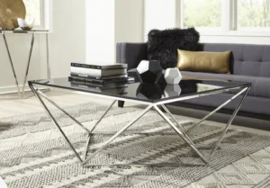 In een moderne woonkamer staat de Salontafel Rookglas, Gerookt Rvs salontafel op een vloerkleed met patroon, gecombineerd met een donkergrijze bank met gouden en zwarte kussens. Decoratieve boeken en vazen sieren de tafel, terwijl grote ramen de ruimte vullen met natuurlijk licht.
