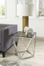 Een moderne woonkamer met een grijze bank, de Aria gerookt glas en gepolijst roestvrijstalen bijzettafel, een gouden en witte tafellamp en een wit decoratief olifantenbeeld bij grote ramen.
