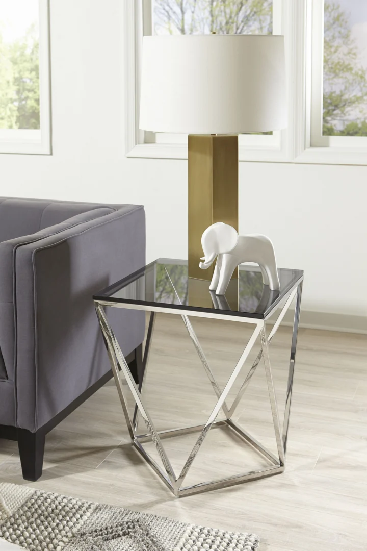 Een moderne woonkamer met een grijze bank, de Aria gerookt glas en gepolijst roestvrijstalen bijzettafel, een gouden en witte tafellamp en een wit decoratief olifantenbeeld bij grote ramen.