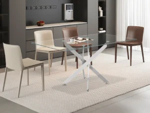 Een moderne eetkamer met de Tafel 'Lasi' 160 cm Glas met chromen poten, vier beige en bruine stoelen, een gestreept vloerkleed, een koffiezetapparaat op het aanrecht, decoratieve planken tegen een donkere muur en zonlicht uit een groot raam.
