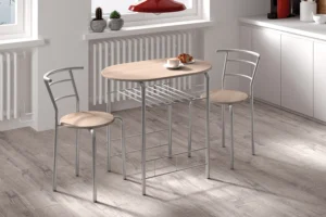 De Tafelset 'Adam' Grijs metaal Decor sonoma heeft een compacte, moderne ovale tafel van licht hout met twee bijpassende stoelen met metalen frame, ideaal voor een lichte keuken. Bovenop de tafel staan een croissant en een kopje; kleine plantjes sieren de vensterbank.