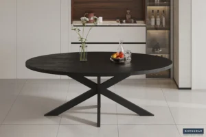 De Tafel 'Alexa' 200 cm Ovaal van zwart mangohout met schuine poten staat op een betegelde vloer. Bovenop staan een glazen vaas met bloemen, een karaf, twee glazen en een fruitschaal. Minimalistische kasten zijn op de achtergrond zichtbaar.