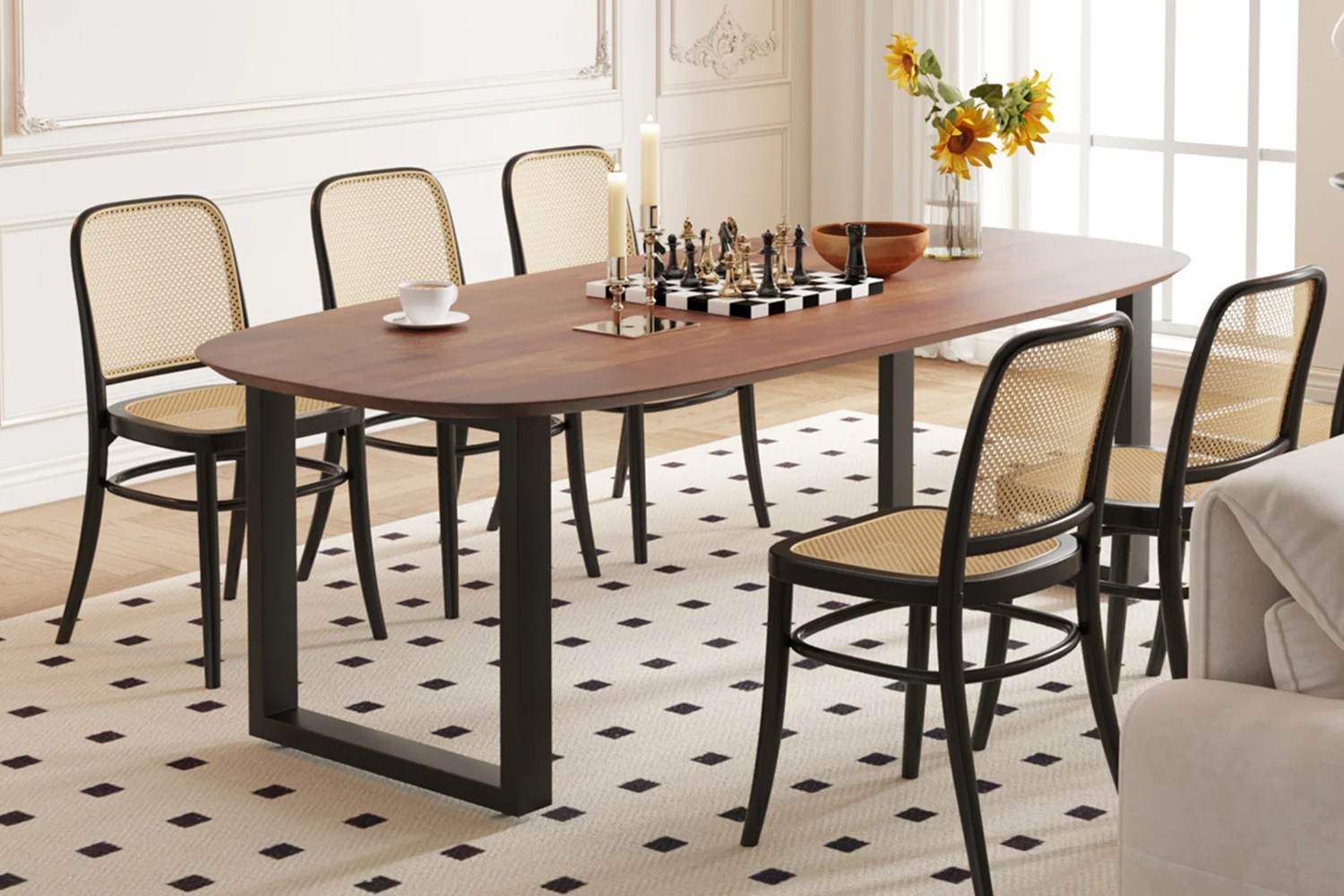 In een moderne eetkamer staat de UITVERKOOP - Tafel 'Tommy' 220x100 cm Deens Ovaal U-Poot in bruin mangohout, een set voor zes personen met stoelen met rieten rugleuning, een schaakspel, zonnebloemen in een vaas, een schaal, kaarsen en een theekopje op een licht gedessineerd vloerkleed. Zonlicht vult de ruimte.