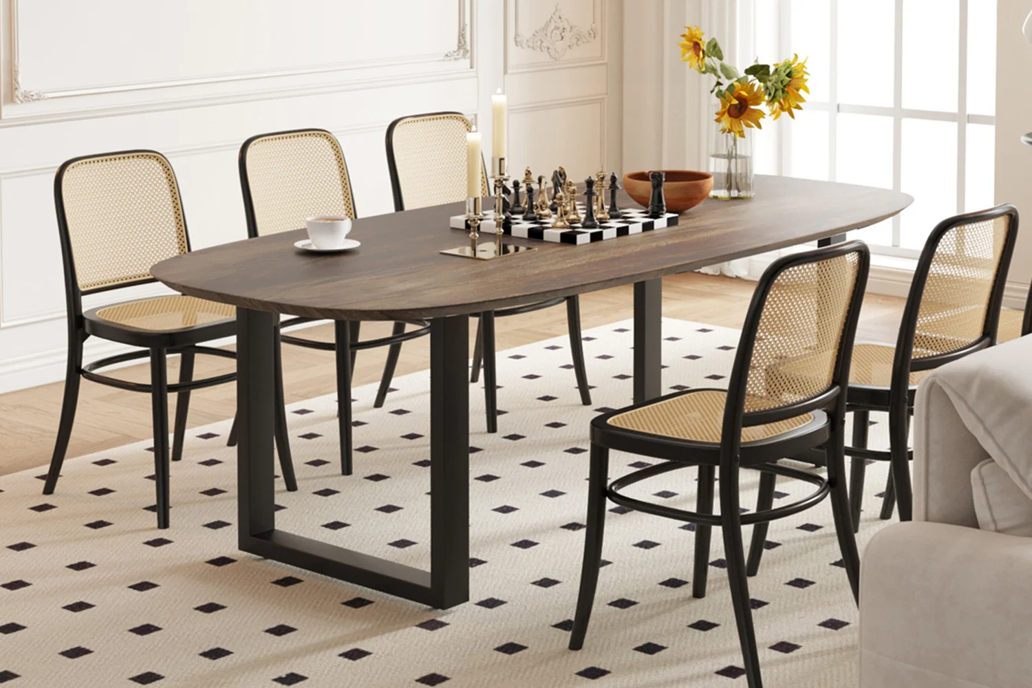 In een moderne eetkamer staan de UITVERKOOP - Tafel 'Tommy' 220x100 cm Deens Ovaal U-Poot Mango hout Tabaco, zes stoelen met rieten rugleuning, een schaakbord, kaarsen, een beker, een schaal en een vaas met zonnebloemen bij een groot raam met daglicht.