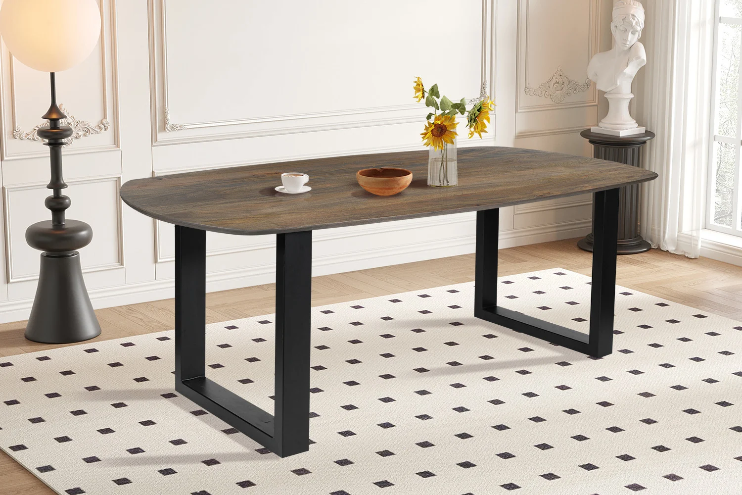 De UITVERKOOP - Tafel 'Tommy' 220x100 cm Deens Ovaal U-Poot Mango hout Tabaco staat op een wit gedessineerd vloerkleed, met daarop zonnebloemen in een vaas, een houten schaal en een witte kop en schotel; op de achtergrond staan een buste en een lamp.