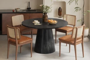 In een moderne, minimale eethoek staat de Tafel 'Tommy' 130 cm Rond Mango hout Zwart, met daarop een vaas met bloemen en een fruitschaal, omringd door vier houten stoelen met rieten rugleuningen en leren zittingen.