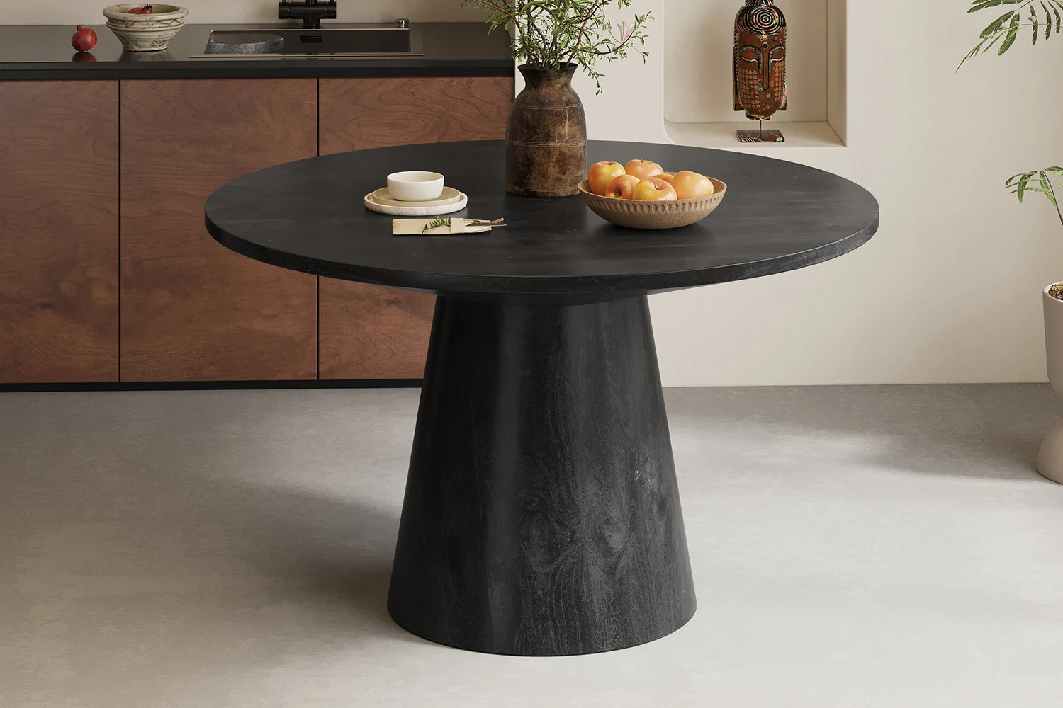 In een moderne eetkamer staat de Tafel 'Tommy' 130 cm Rond Mango hout Zwart, een ronde zwarte tafel met een breed voetstuk. Decor bestaat uit een bruine vaas met groen, sinaasappels in een schaal, een kopje en eetstokjes op een bord. Minimalistische stijl voert de boventoon.