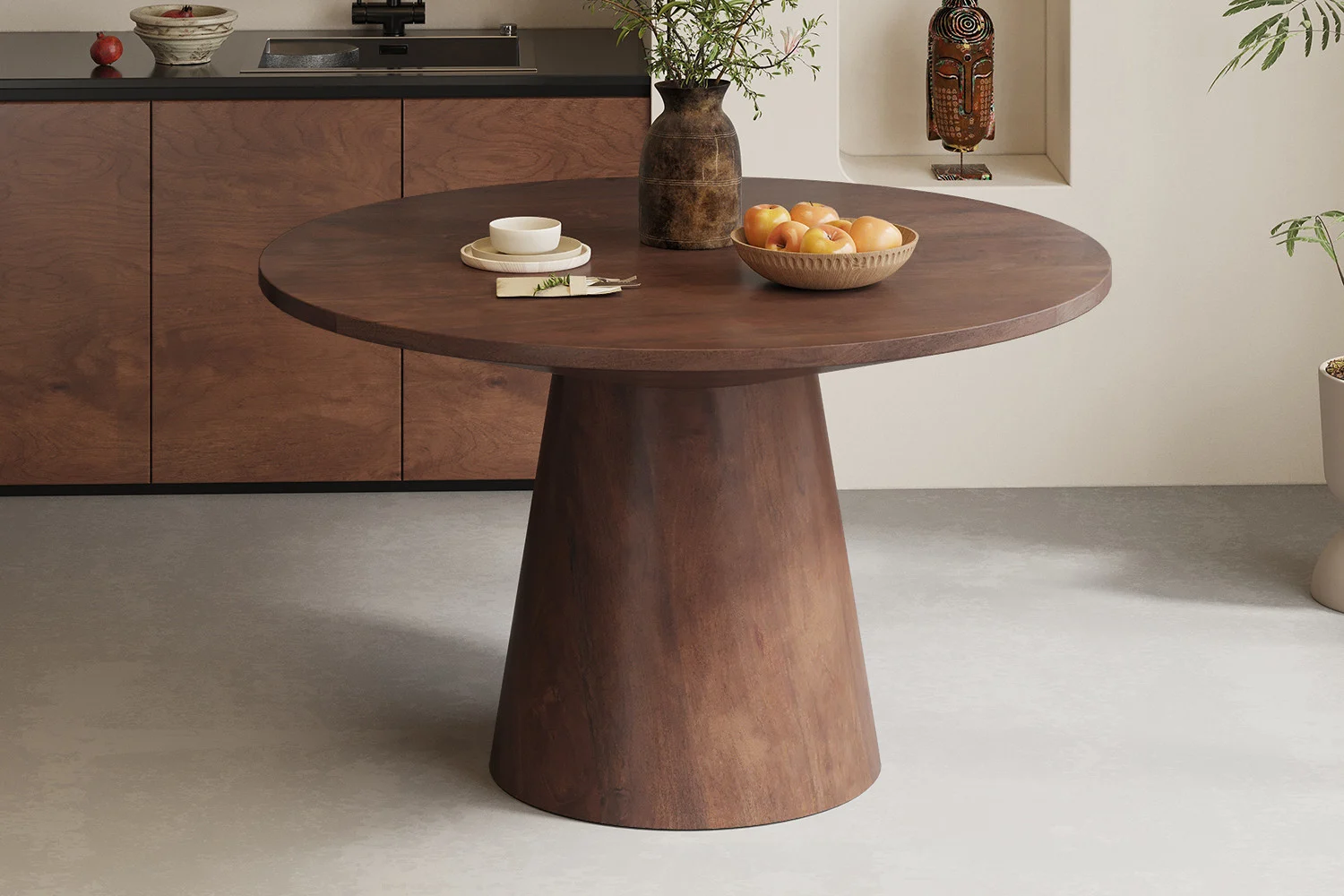 De Tafel 'Tommy' 130 cm Rond Mango hout Bruin is te zien in een moderne keuken, gestyled met een bruine vaas met groen, een schaal met appels en boeken erop, met bruine kasten op de achtergrond.