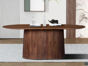 De Tafel 'Miguel' 200 cm Ovaal van lichtbruin mangohout heeft een brede cilindrische basis en staat op een tapijt met structuur, met daarop glaswerk, een waterkaraf, een glas en een zwarte decoratieve neushoorn te midden van een minimalistisch decor.