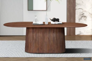 De Tafel 'Miguel' 230 cm Ovaal Mango hout Lichtbruin staat op een grijs vloerkleed met textuur, een ovaal lichtbruin houten blad, een gebogen gegroefd onderstel en decor waaronder een glazen karaf, twee glazen, boeken en een zwart neushoornbeeld.