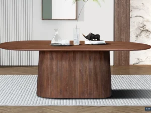 De Tafel 'Miguel' 230 cm Ovaal Mango hout Lichtbruin staat op een grijs vloerkleed met textuur, een ovaal lichtbruin houten blad, een gebogen gegroefd onderstel en decor waaronder een glazen karaf, twee glazen, boeken en een zwart neushoornbeeld.