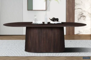 In een moderne eetkamer staat de Tafel 'Miguel' 230 cm Ovaal Mango hout Donkerbruin met een gebogen voet. Bovenop staan een glazen karaf, twee glazen, een boek en een zwart neushoornbeeldje, allemaal gerangschikt op een gestreept tapijt in een minimalistische ruimte.