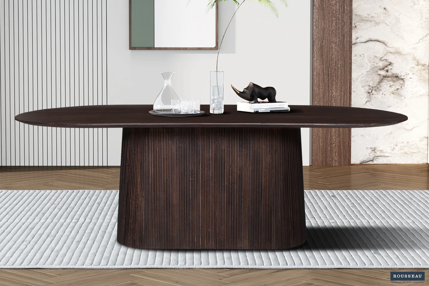 In een moderne eetkamer staat de Tafel 'Miguel' 230 cm Ovaal Mango hout Donkerbruin met een gebogen voet. Bovenop staan een glazen karaf, twee glazen, een boek en een zwart neushoornbeeldje, allemaal gerangschikt op een gestreept tapijt in een minimalistische ruimte.