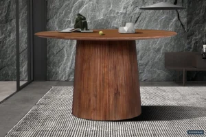 De Tafel 'Miguel' 130 cm Rond Mango hout Lichtbruin staat op een zwart-wit gedessineerd tapijt, versierd met decoratieve items en boeken, tegen een grijze stenen muur.