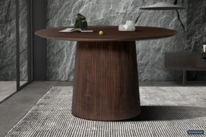 De Tafel 'Miguel' 130 cm Rond Mango hout Donkerbruin met een geribbelde cilindrische voet staat op een gestreept kleed bij een grijze stenen muur met structuur, waarop decoratieve objecten en een kleine groene bal staan.