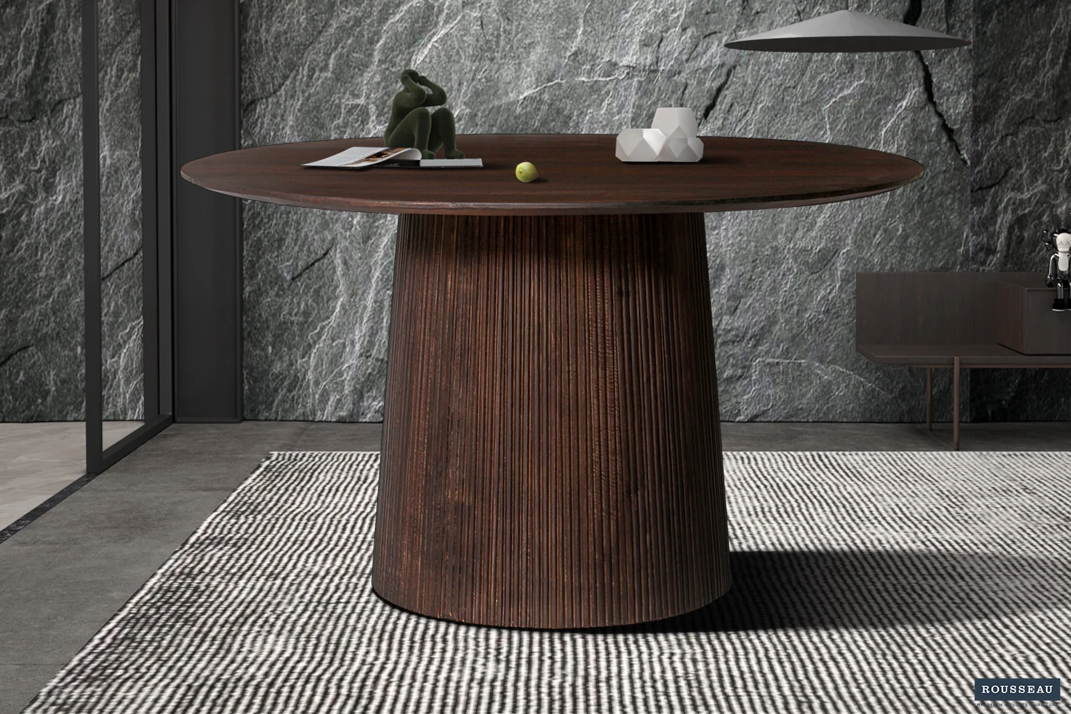 De Tafel 'Miguel' 130 cm Rond Mango hout Donkerbruin met een geribbelde cilindrische voet staat op een gestreept kleed bij een grijze stenen muur met structuur, waarop decoratieve objecten en een kleine groene bal staan.