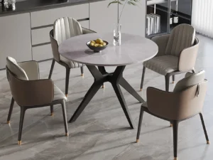 De Tafel 'Apollo' ronde tafel van 110 cm heeft een glanzend Italiaans grijs steenachtig oppervlak en zwarte poten, omringd door vier gecapitonneerde taupe stoelen. Op de tafel staan een glazen vaas met een plant en een fruitschaal; de kamer heeft grijze kasten en betegelde vloeren.