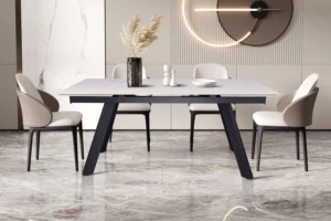 In een moderne eetkamer staan de Uitschuifbare tafel 'Hera' 160-240 cm Rechthoekig Steen Wit, vier stoelen met kussens, een vaas met gedroogd pampagras, twee kandelaars en een wandklok op marmeren vloeren met decoratieve wandpanelen.