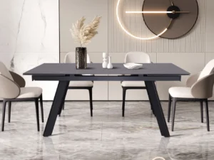 Een moderne eetkamer met de Uitschuifbare tafel 'Hera' 160-240 cm Rechthoekig Steen Zwart, vier beige stoelen, een vaas met gedroogd pampagras, keramisch decor, marmeren vloeren en een wandklok met cirkelvormig licht.