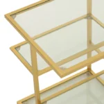 Een close-up van de Bijzettafel ijzer/glas goudkleurig toont het moderne goudkleurige metalen frame en de drie glazen legplanken, die een geometrisch ontwerp met transparante oppervlakken hebben.
