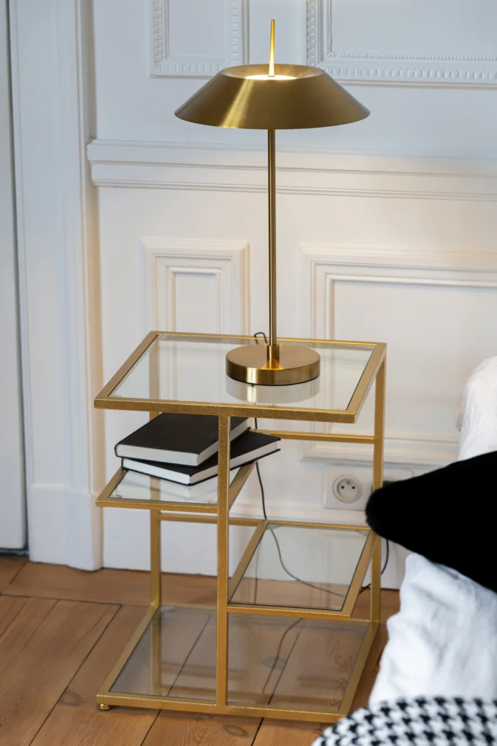 Een moderne Bijzettafel ijzer/glas goudkleurig houdt een bijpassende gouden lamp en twee gestapelde boeken vast; tegen een wit gelambriseerde muur, met een glimp van een bed en houten vloer op de achtergrond.