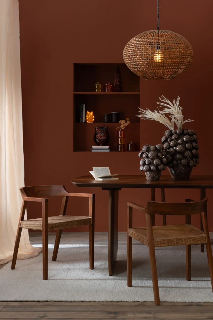 Een moderne eethoek met de Eettafel Acaciahout Bruin ronde tafel met twee houten stoelen, sculpturale vazen met gedroogde planten, een rieten hanglamp en ingebouwde planken, allemaal tegen een warmbruine muur die zacht wordt verlicht door natuurlijk licht.
